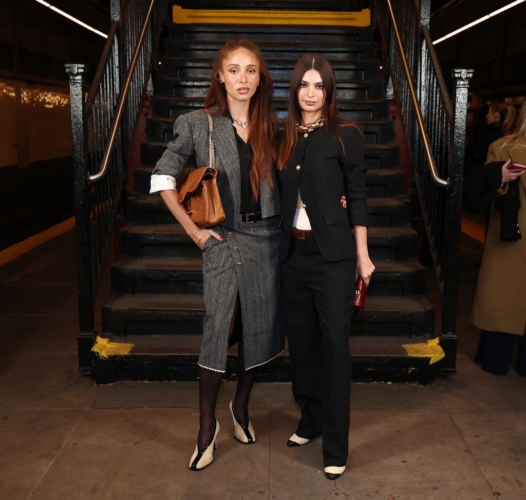 Adwoa Aboah and Emily Ratajkowski pose in the subway. Adwoa wears a grey skirt suit with sheer black tights, monochrome heels and a large brown handbag over her shoulder. Her red hair is worn long and loose.