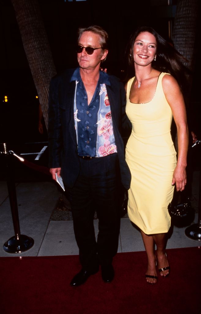 Michael Douglas, wearing a black suit over a floral pattern shirt and sunglasses, and his partner, British actress Catherine Zeta-Jones, who wears a sleeveless yellow dress, attend the Beverly Hills premiere of 'The Muse' at the Academy Theater in Beverly Hills, California, 16th August 1999