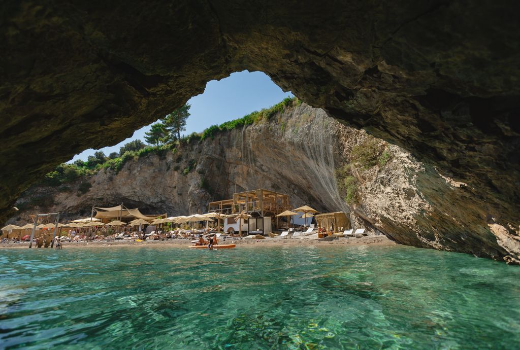 Une vue de l'intérieur d'une grotte marine sur une plage de Lustica Bay, Monténégro. L'entrée de la grotte encadre une plage étroite avec des chaises longues et des parapluies, et des gens se détendant par l'eau turquoise. 