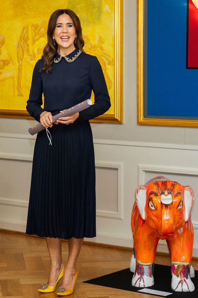La reine Mary souriante en robe bleu marine et talons jaunes tenant un parchemin