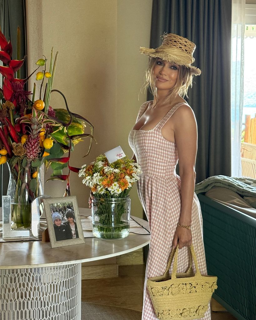 In this warm illuminated inner photo, Jennifer Lopez is graceful next to a round table that is adorned with lively flower arrangements and a framed photo of two children. She wears a soft rosa white Gingham dress in the milk maid style, which hugs her figure and gently torches on the floor. Your hair is designed in relaxed waves and partly hidden under a Raffia hat in Raffia. On her arm, she wears a straw-containing Raffia shopping bag with complicated crocheting flower crochet. The ambience is airy and summer, with natural light streaming from a window in the background and emphasizing the coastal setting beyond the room. Your expression is calm and exudes a casual elegance.