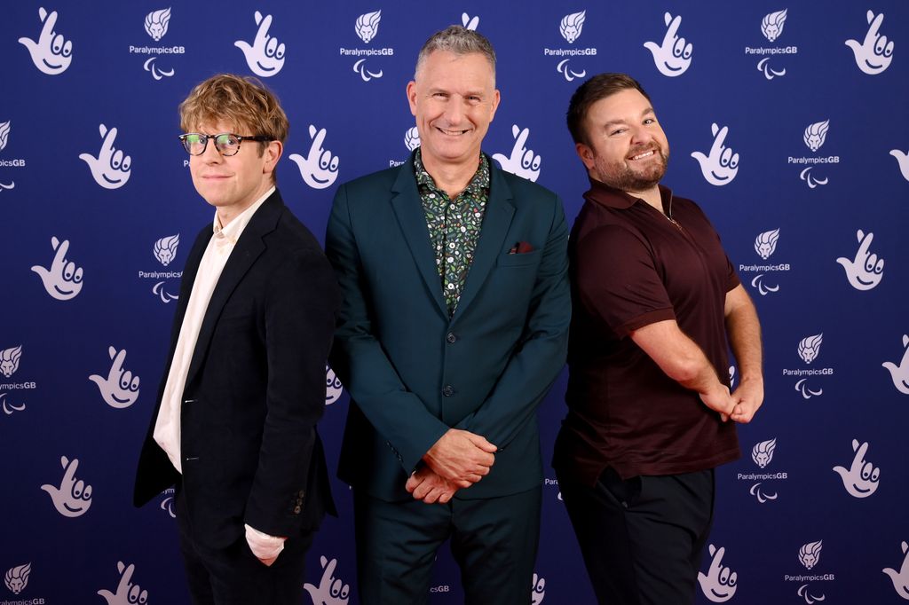 Josh Widdicombe, Adam Hills and Alex Brooker in front of national lottery promotional board posing for a photo