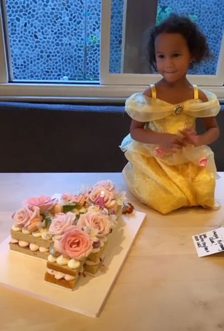 a little girl kneels on the floor wearing a yellow dress which looks like the character belles dress from the disney film beauty and the best and on the floor beside her is a pretty sponge cake filled with cream and topped with pink flowers