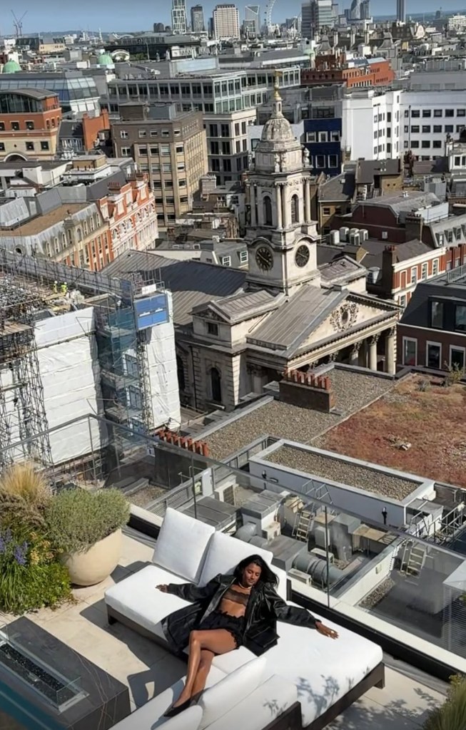 Bridgerton actress Simone Ashley sits on a rooftop - the London skyline visible behind her - wearing an all-black ensemble, sprawled out on a white sofa. 
