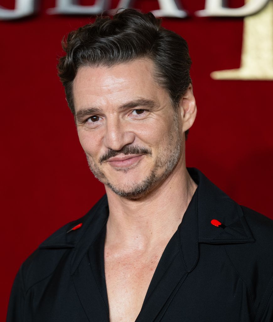 Pedro Pascal in a low plunging black shirt with red detailing, sporting a moustache and coiffed hair. 