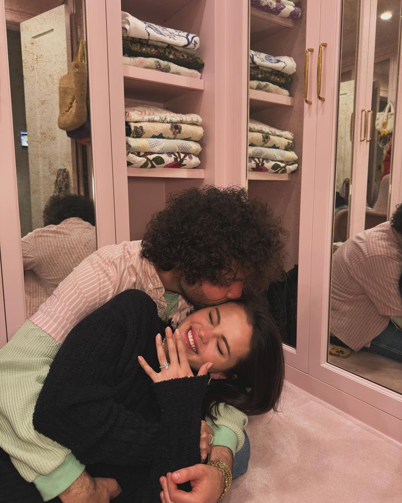 Selena Gomez and Benny Blanco share a kiss in their wardrobe while Selena shows off her engagement ring. Selena wears a black long sleeve jumper and Benny wears a pink and mint green top.