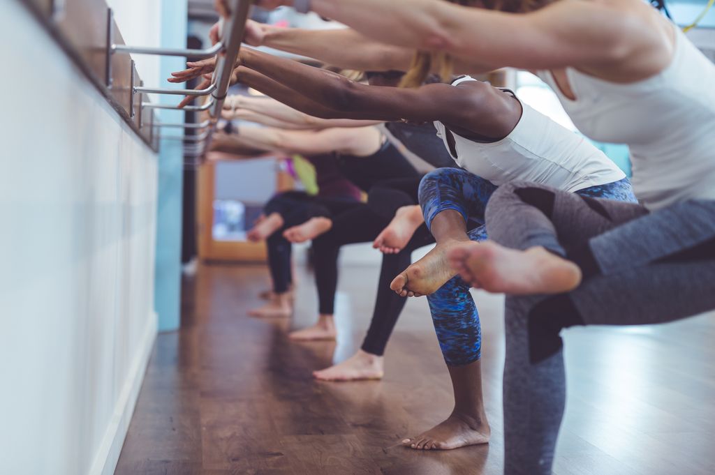 group of young women do a barre workout together at a modern gym. They are facing the wall, gripping the bar with both hands, and doing repetitions of squats, with one foot crossed over the opposite thigh.
