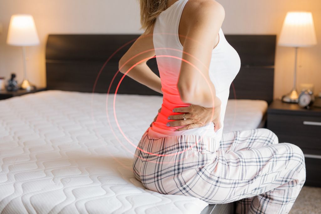 A woman sitting on the edge of a bed holding her lower back, with a red circular graphic overlay highlighting a concentrated area of inflammation or pain.