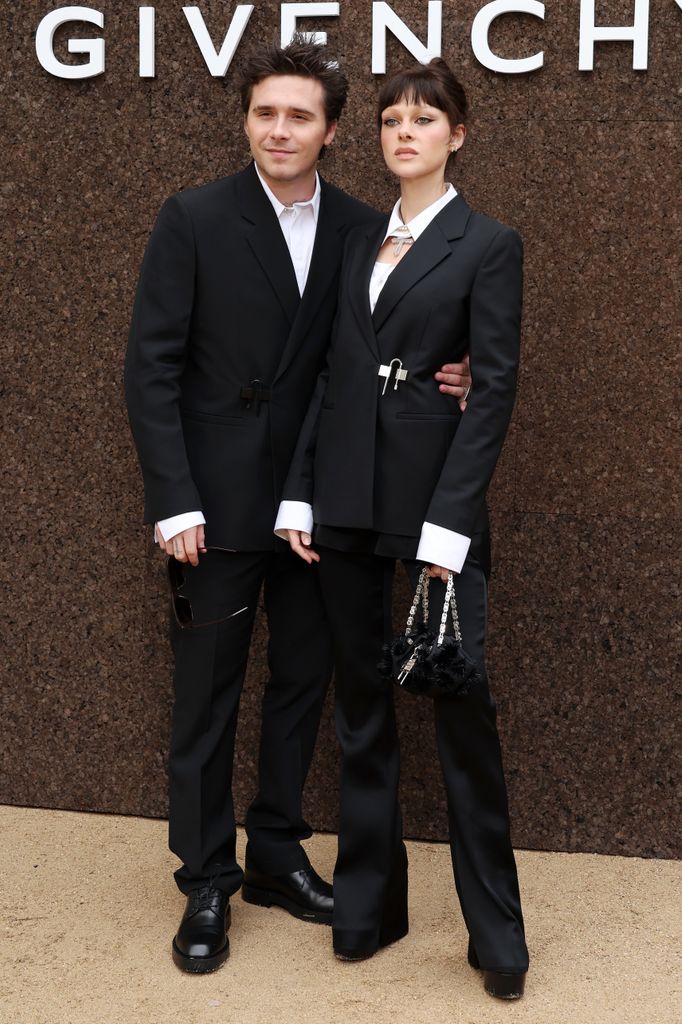 This image features two individuals standing together in front of a textured brown wall with the large word "GIVENCHY" prominently displayed above them. They are both dressed in elegant, coordinated black outfits.  The person on the left is wearing a tailored black suit with a white dress shirt underneath. The suit jacket has a slightly modern cut with a cinched waist detail. They are also wearing black dress shoes, and they hold a pair of sunglasses in one hand. Their hair is styled in a slightly messy, voluminous look, and they are smiling subtly.  The person on the right is also wearing a stylish black suit but with a more fashion-forward design. The suit jacket is cinched at the waist with a decorative clip, creating a sculpted silhouette. Underneath, a white collared shirt is visible. They pair the suit with wide-legged black pants and platform black shoes. They are accessorizing with a small, structured black handbag that features intricate black floral details and a silver chain handle. Their hair is styled in a chic updo with bangs, and their makeup includes defined eyes and lips, giving a polished and striking look.  Both individuals are standing close together, with the person on the left placing their arm around the other in a supportive gesture. The overall tone of the image conveys high-fashion elegance and sophistication, likely at a designer event or show.