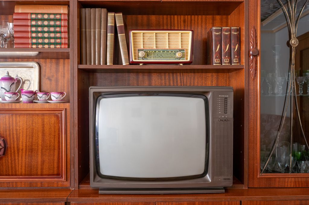 Old television and radio sit on a wooden shelf in a 1980s living room, surrounded by books and a tea set, showcasing the retro home interiors of the era