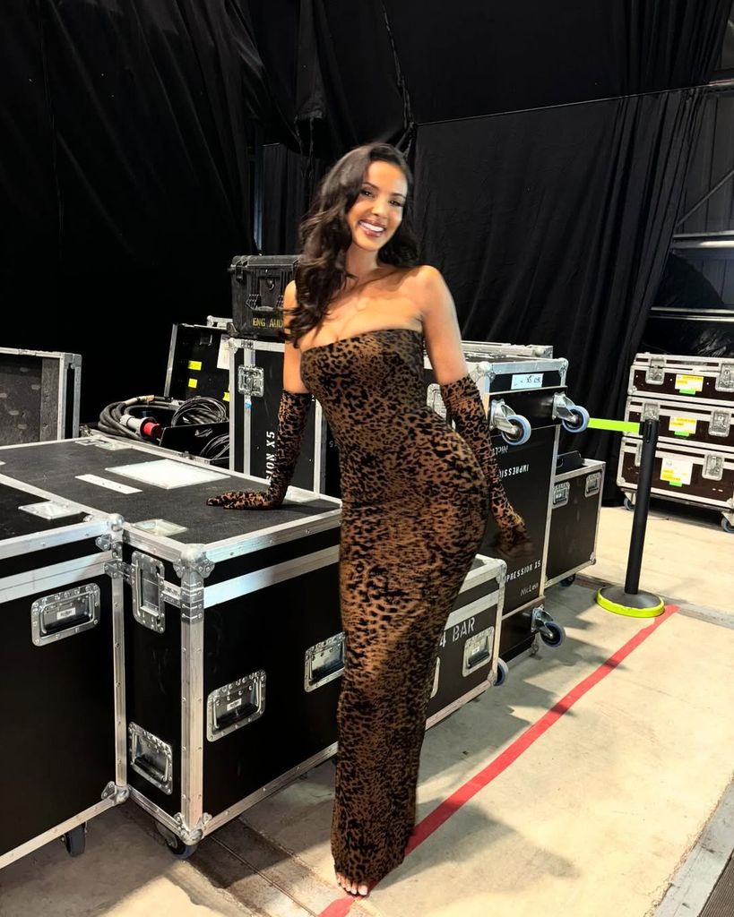 Maya smiles brightly in a backstage setting, wearing a fitted leopard-print gown with long gloves. Equipment cases and cables surround her, adding contrast to her elegant look.