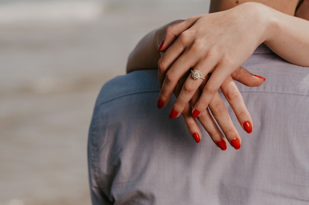 a woman wrapping arms around man's shoulders with red nails and engagement ring on display