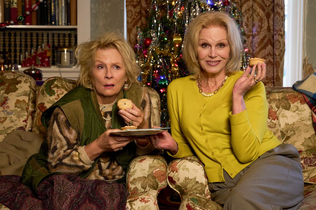two women eating mince pies sitting on sofa in front of christmas tree