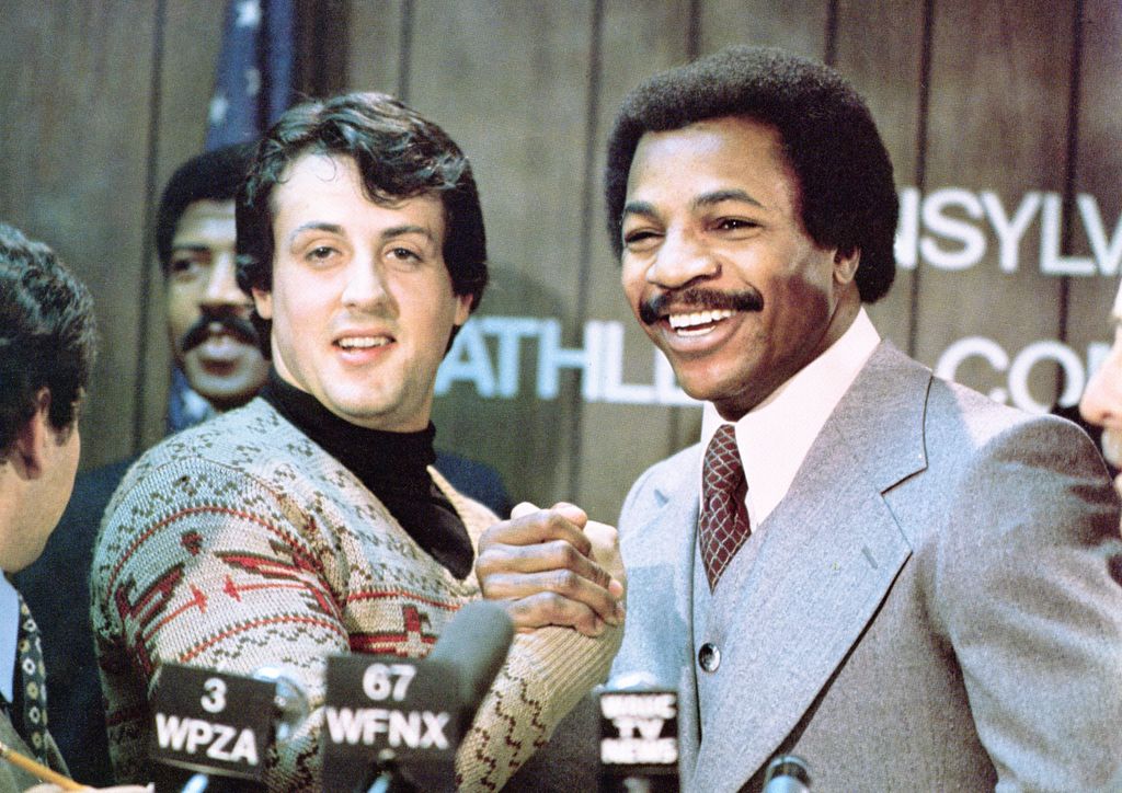Sylvester Stallone (L) and Carl Weathers grip hands and smile together during a press conference in a still from the film, 'Rocky,' directed by John G. Avildsen, 1976