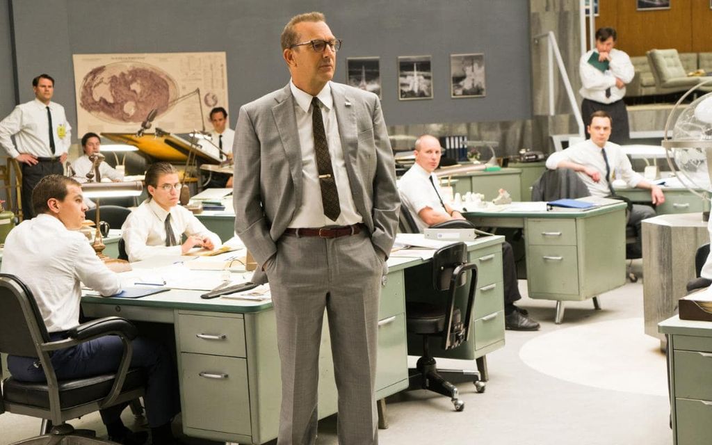 Kevin Costner in Hidden Figures; he stands in a suit while men sit at desks behind him.