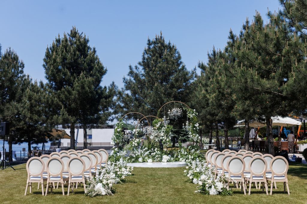 Luxurious outdoor wedding reception is decorated with fresh white flowers and evergreens. Three arches are on the white podium in the middle. Clear blue sky and green pines are on the background.