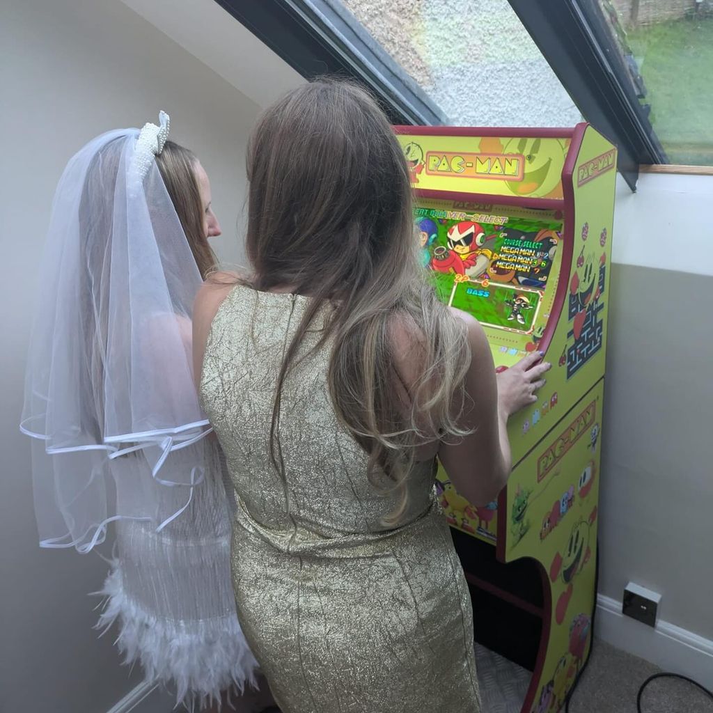 A bride and a woman in a gold dress playing on a pacman machine 