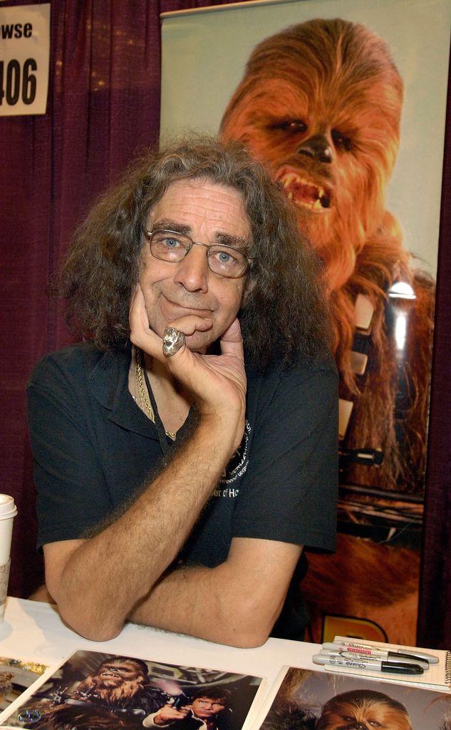 Peter Mayhew est assis devant une bannière représentant Chewbacca tenant sa tête dans une main.