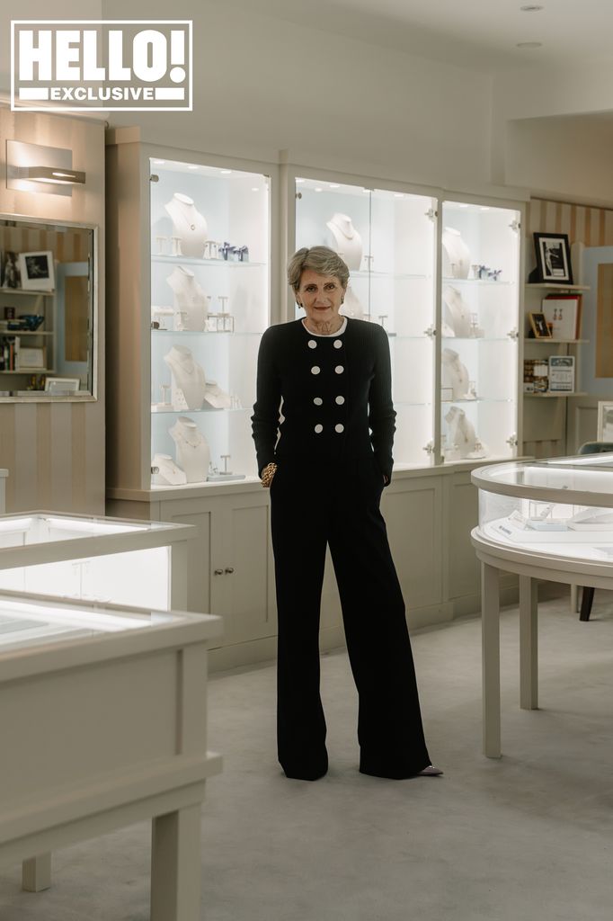 A woman wearing black trousers and a black cardigan with her hands in her pocket, posing in the middle of a jewellery shop