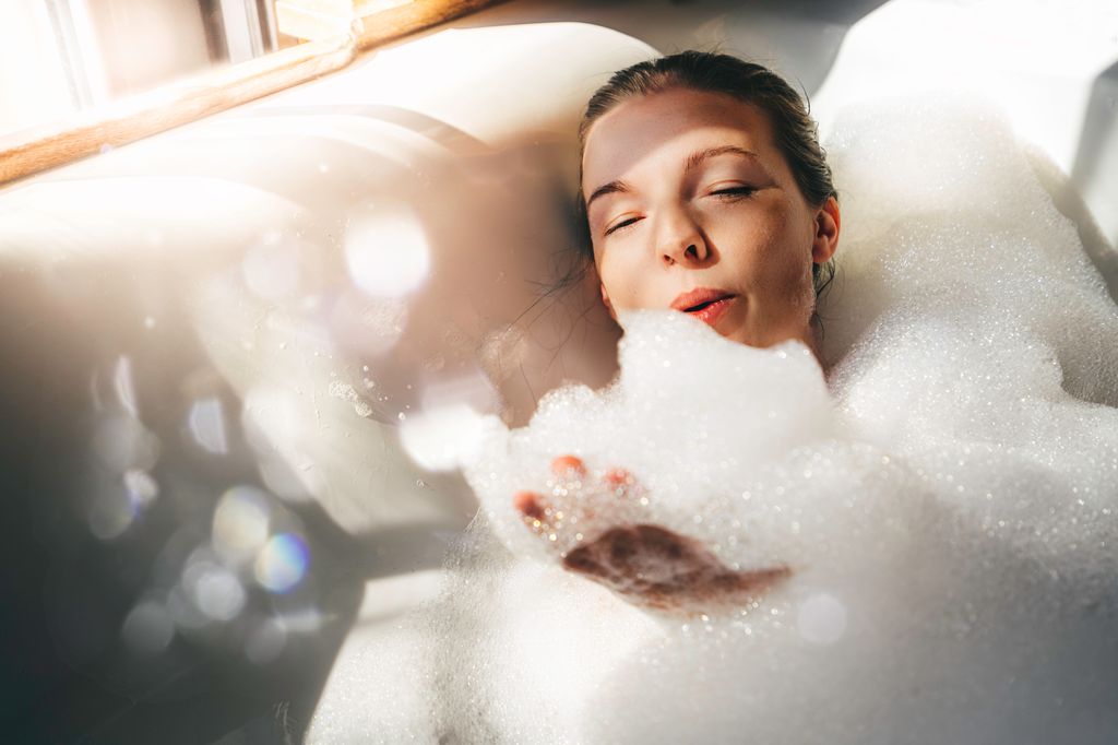 Beautiful woman relaxing in hot tub in a hotel room. She is taking time for herself in a bubble bath, hair tucked up in a bun, sun light through curtains. The female is serene and happy. Concepts of self-love, self-care, mindful and conscious attitude to the health
