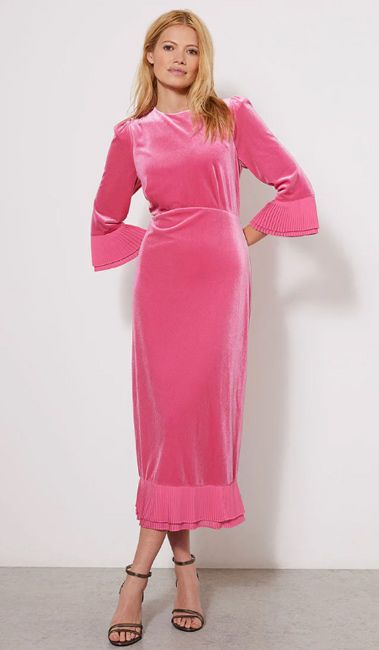 a model wearing a long pink fitted velvet dress with pleats at the hemline and sleeves with her hands behind her back