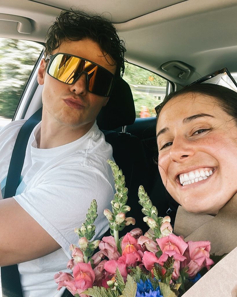 Dominik Fischnaller fait la moue en portant des lunettes de soleil et en conduisant une voiture, avec Emily Sweeney assise sur le siège passager, souriante et portant des fleurs roses.