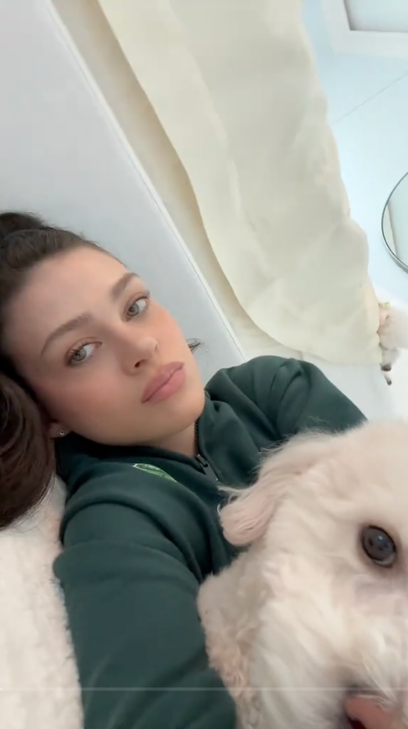 Nicola Peltz lounges on a white sofa in a forest-green hoodie, holding a fluffy white dog close. Her relaxed expression complements the soft, natural light.