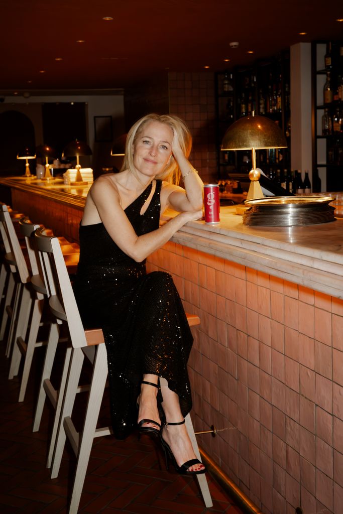 Gillian Anderson rocks a black bedazzled dress as she sits on a bar stool next to a can of her G Spot soft drink