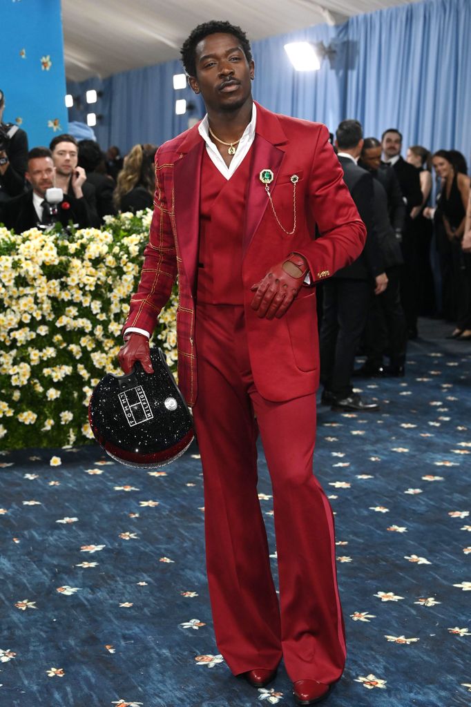 Damson Idris wears a red three piece suit with flared trousers and carries a bedazzled motorcycle helmet.