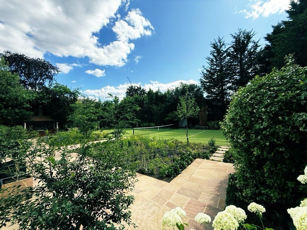 Photo of Ricky Gervais' tennis court in his garden, surrounded by flowers, plants and trees