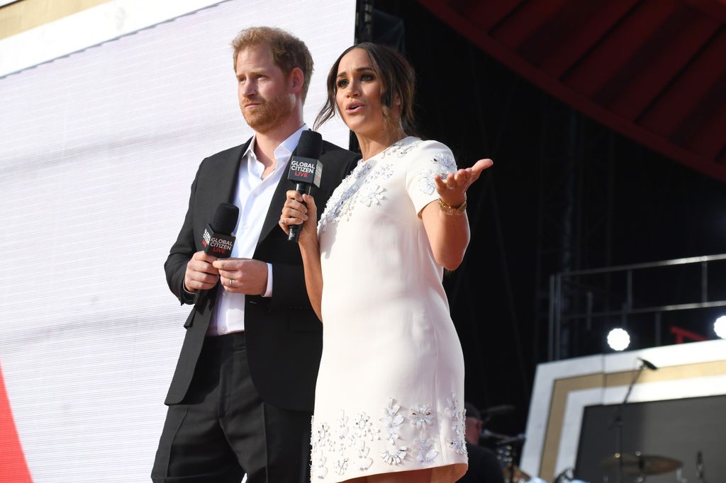 Meghan in white dress talking into mic beside harry