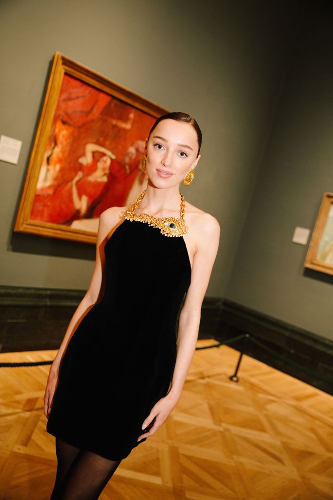 Actress Phoebe Dynevor stands confidently between two grand neoclassical columns, exiting an opulent building. She wears a sleek, floor-length black leather coat over a short, velvet black dress adorned with a striking gold necklace detail. Her legs are bare beneath sheer black stockings, and she finishes the look with classic black stiletto pumps. Her hair is pulled back in a polished bun, and she wears minimal makeup, accentuating her natural features. The setting is warm and intimate, with glowing interior lighting casting a soft, elegant ambiance.