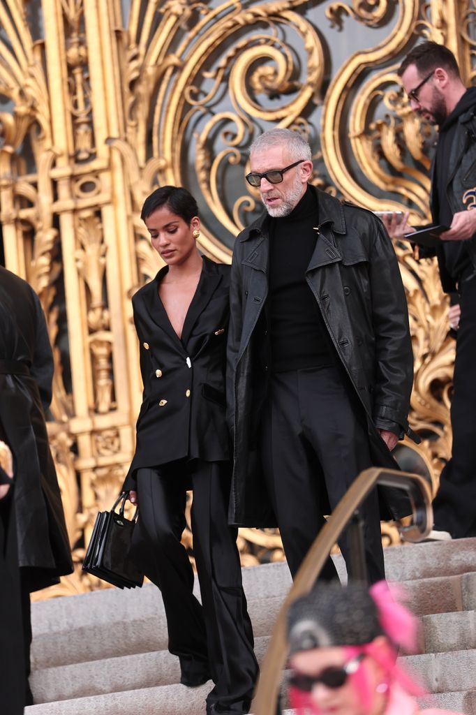 Narah Baptisa and Vincent Cassell walk down a set of steps wearing all black ensembles. 