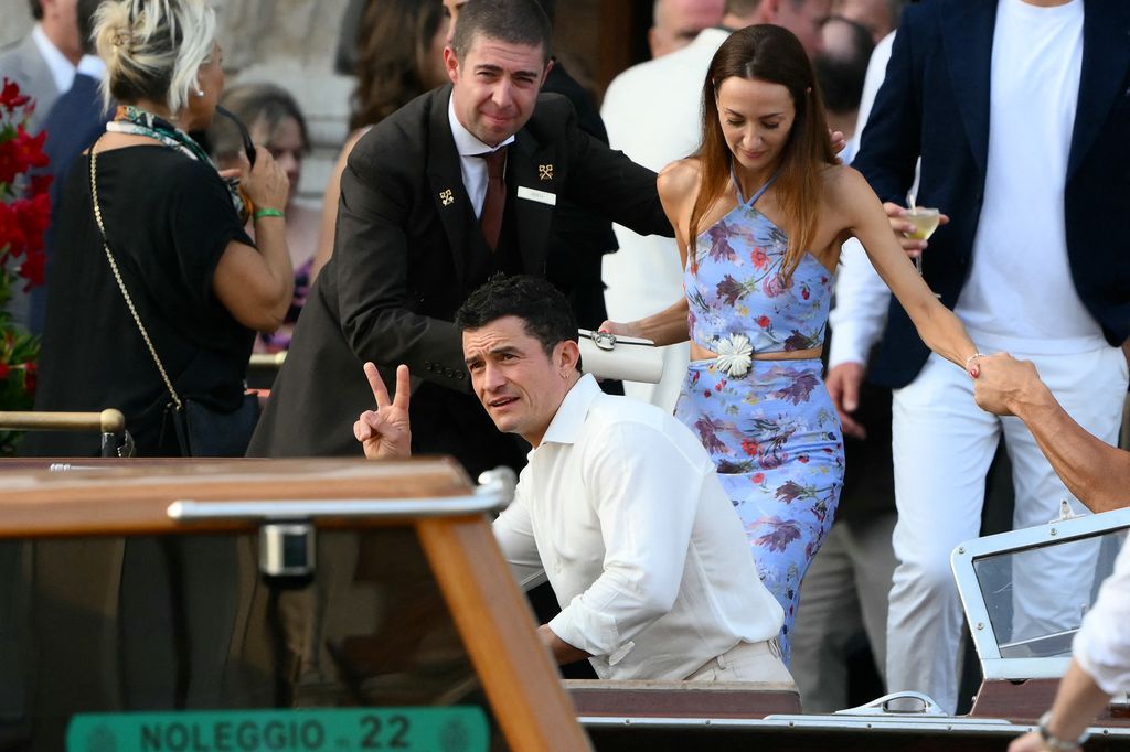 Orlando Bloom flashes a peace sign while wearing a white shirt, joined by a guest in a floral two-piece halterneck outfit, arriving by boat in Venice.