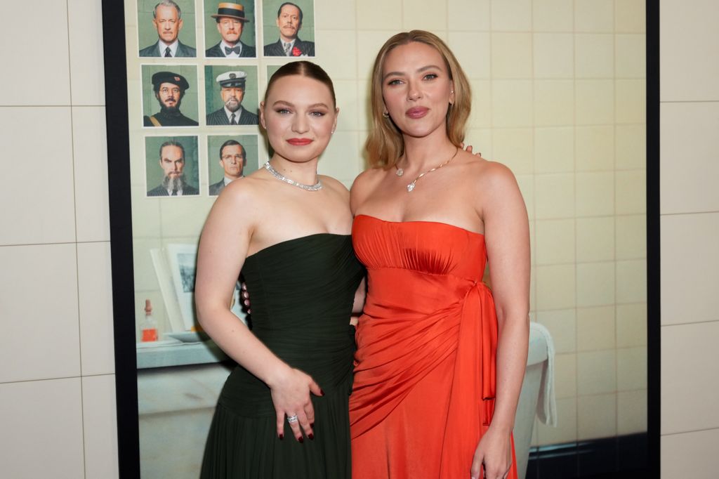 Mia Threapleton and Scarlett Johansson pose together at The Phoenician Scheme premiere in New York, showcasing strapless gowns and dazzling diamond jewellery against a vintage character portrait backdrop.
