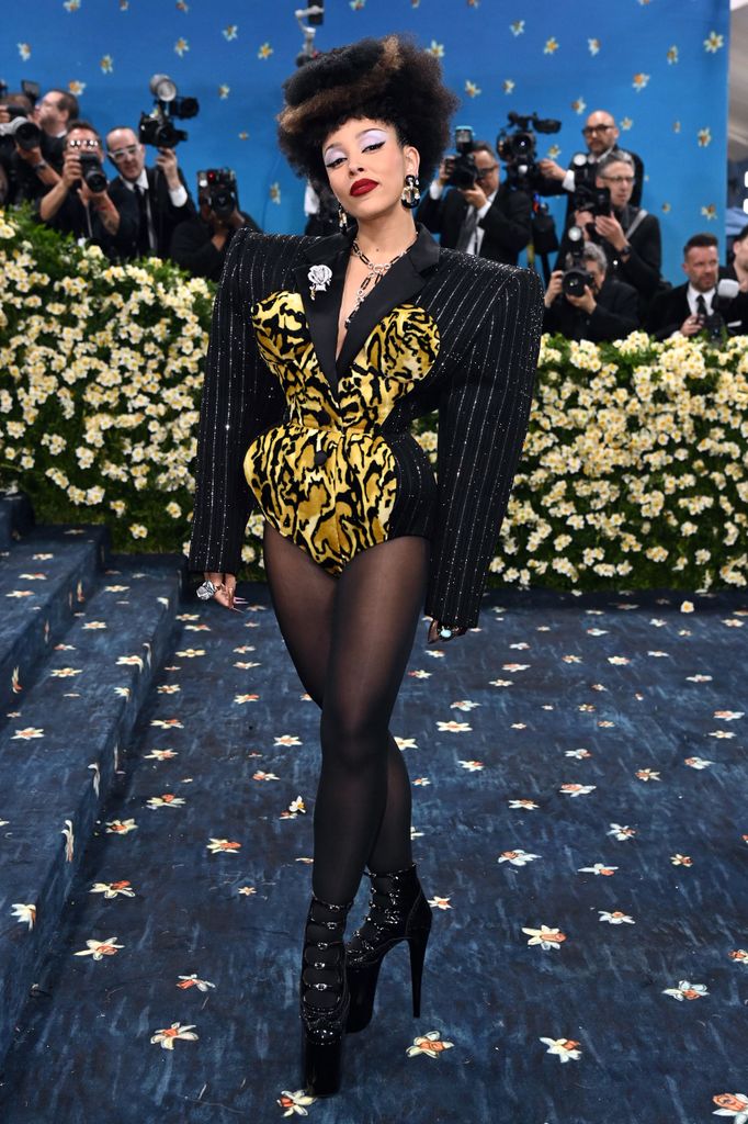 Doja Cat wears a pinstripe blazer playsuit with large shoulder pads and leopard print bodice detailing. She wears platform boots and a furry brown hat.