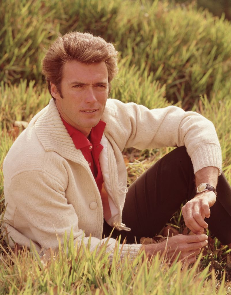Portrait of American actor and director Clint Eastwood sitting in a field, with his leg bent and his elbow resting on his knee, 1960s. Eastwood is wearing a white cardigan over a red shirt