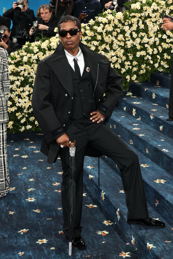A$AP Rocky wears a black suit, white shirt and tie, sunglasses and an umbrella embellished with rhinestones.