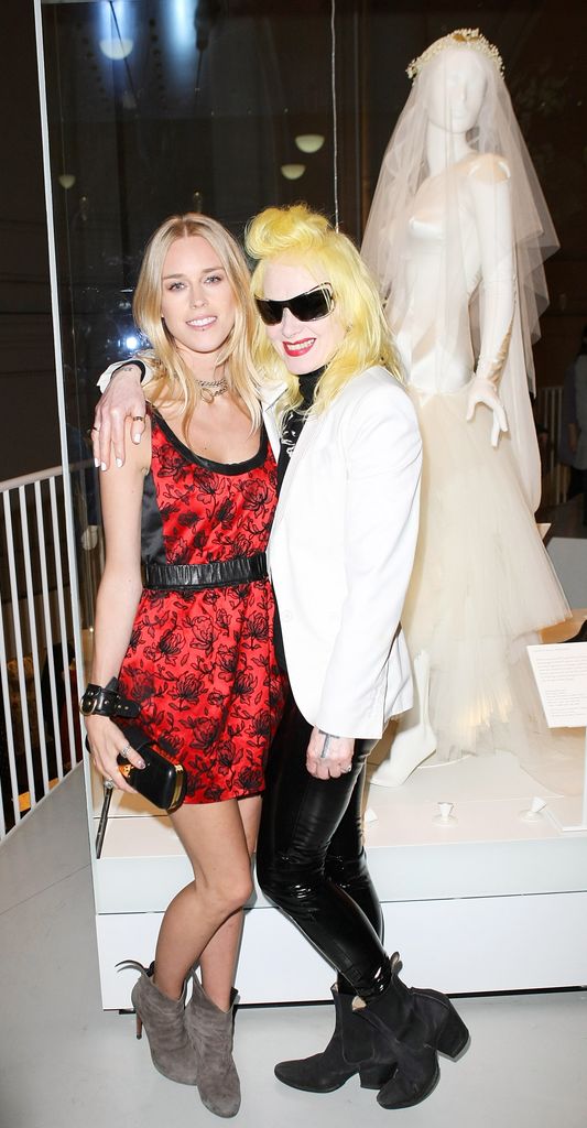 Mme Mary Charteris, vêtue d'une minijupe rouge et de bottines marron, embrasse Pam Hogg, qui porte des lunettes de soleil noires, un blazer blanc et des leggings en latex. Ils se tiennent devant la robe de mariée de Mary sur un mannequin du V&A Museum