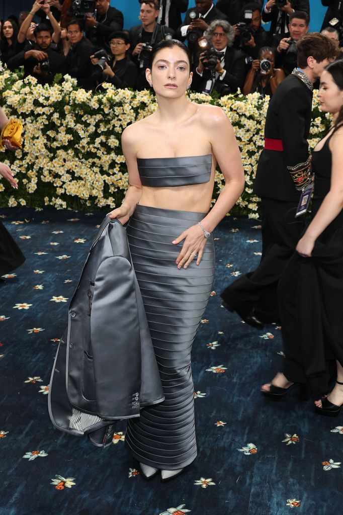 Lorde wears a metallic grey pleated strapless crop top with a matching maxi skirt and blazer.