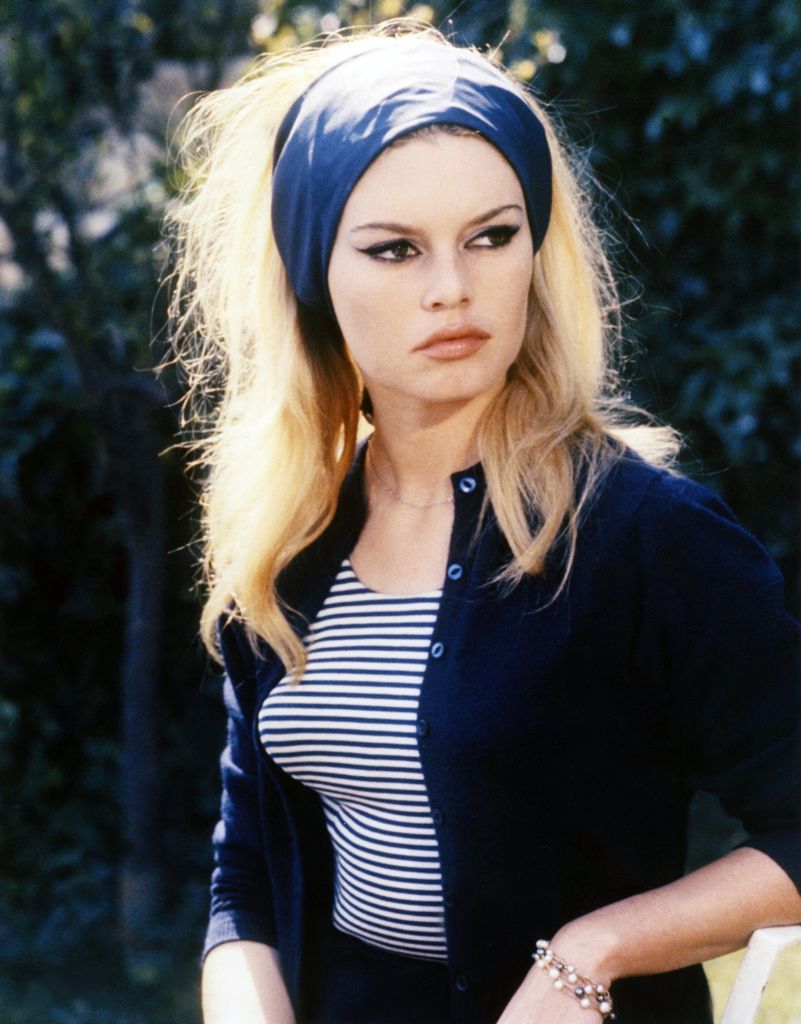 Brigitte Bardot wears a navy headband, blue cardigan and blue and white striped top. Her hair is blonde and bouffant and she has her signature thick black eyeliner on her eyes.