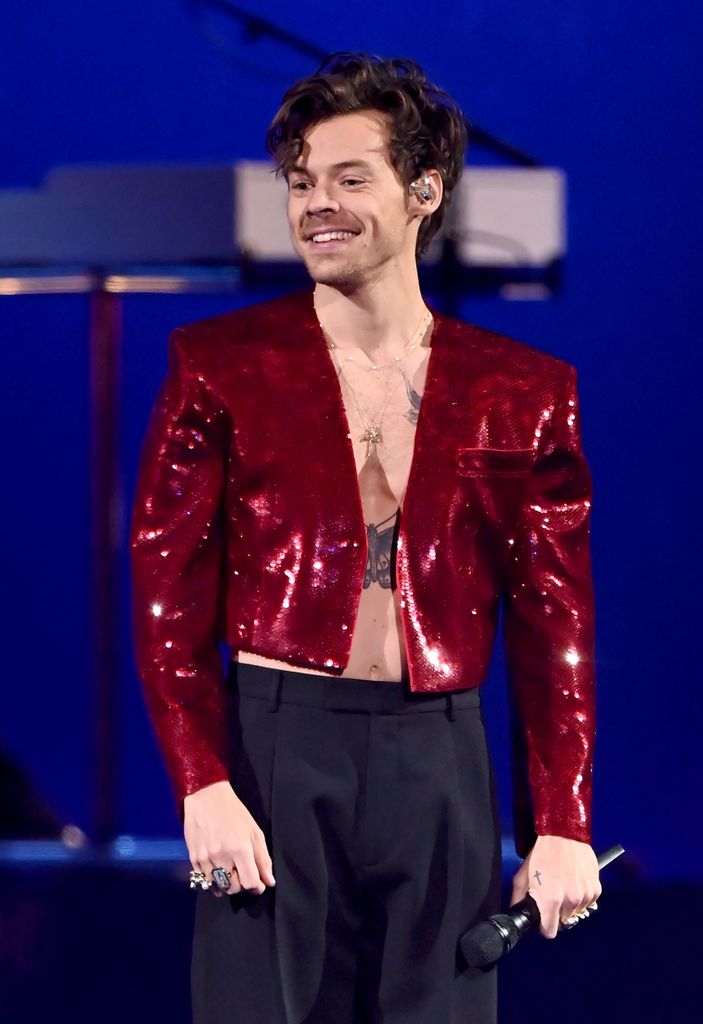 Harry Styles performs on stage in a sparkly red jacket while holding a microphone.