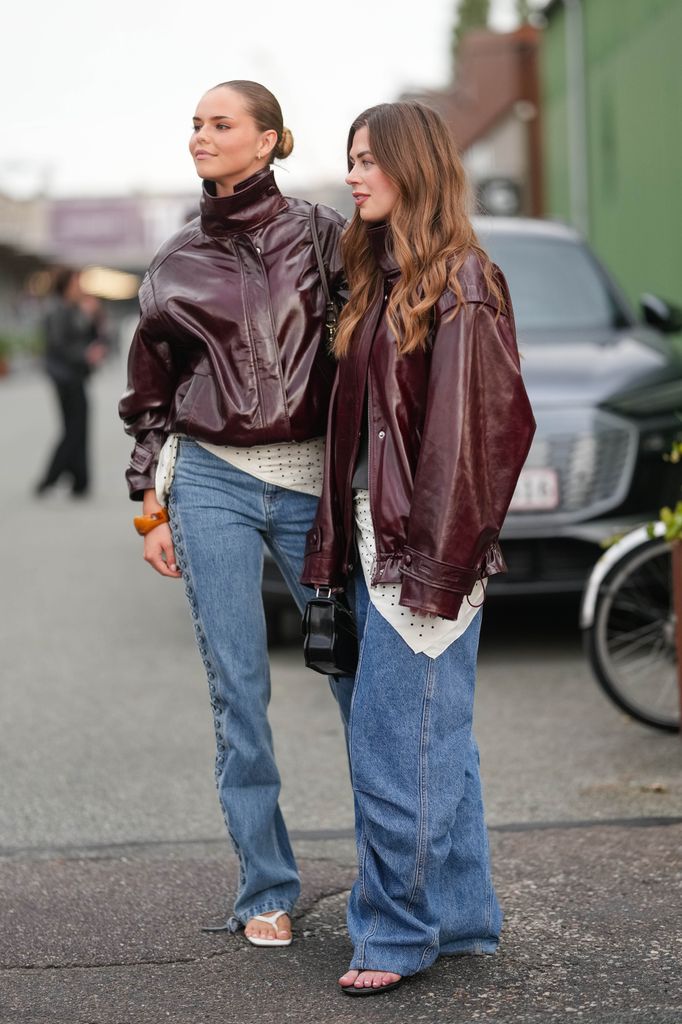  A guest (L) wears a high-neck, dark burgundy leather jacket with a glossy finish, featuring a front zipper and gathered cuffs. Underneath, a white top with black polka dots is visible. Blue denim jeans with a straight-leg cut and button detailing along the side seams are paired with white thong sandals.