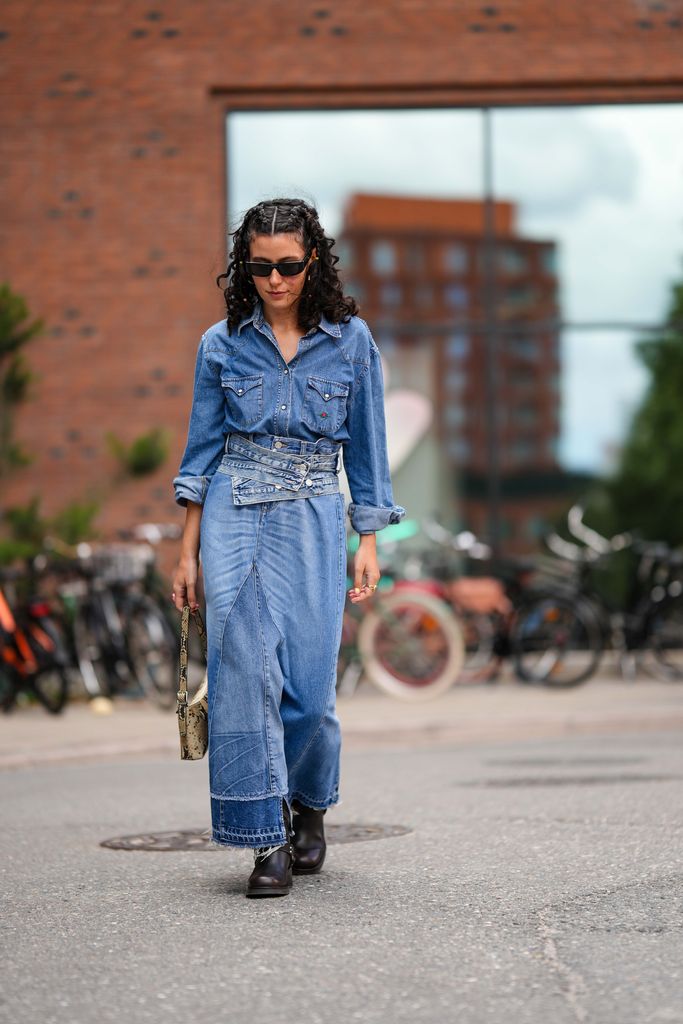 A guest wears black sunglasses from Prada, gold earrings, a blue denim shirt, a blue faded denim ripped long skirt, a blue denim nailed / studded belt, a beige and black snake print pattern shiny leather handbag, black shiny leather ankle boots , outside Lovechild 1979 , during the Copenhagen Fashion Week Spring/Summer 2024 on August 08, 2023 in Copenhagen, Denmark. (Photo by Edward Berthelot/Getty Images)