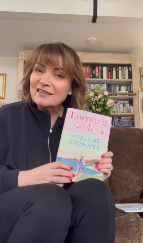 Lorraine holds up her novel while sat on sofa in her living room