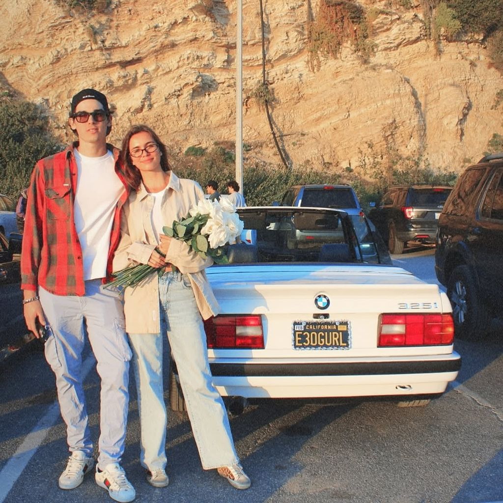 Paris Brosnan and his girlfriend posing in front of white BMW