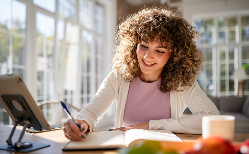 A cheerful woman sits at a desk writing in her journal amidst natural sunlight in an airy, modern home. This setting evokes warmth, focus, and inspiration, ideal for moments of self-reflection.