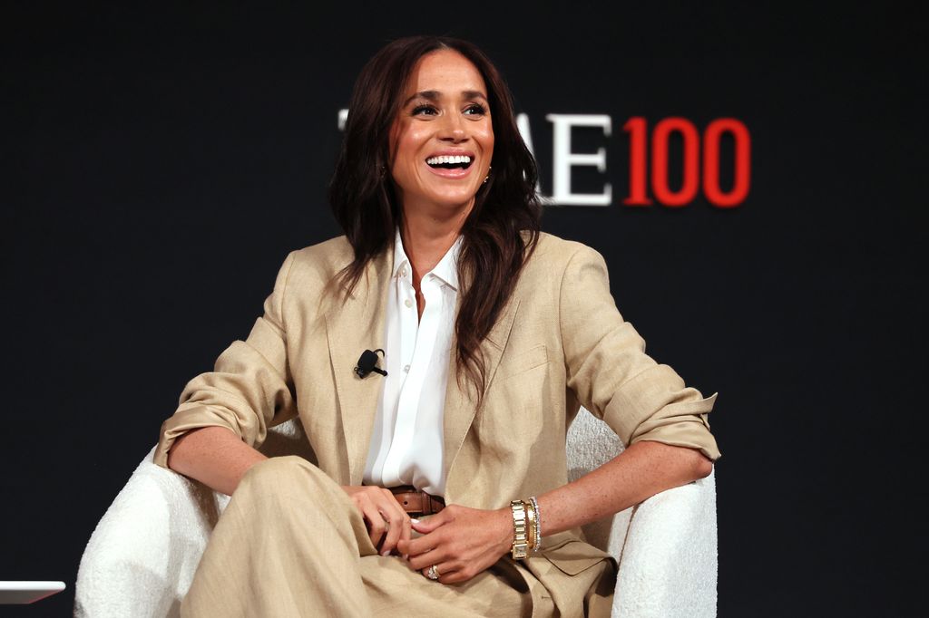 Meghan Markle wearing a linen cream suit smiling while giving a speech