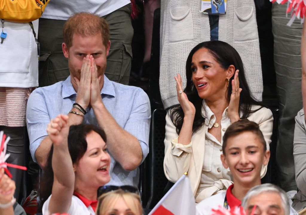 Prince Harry covering his mouth as Meghan Markle applauds next to him