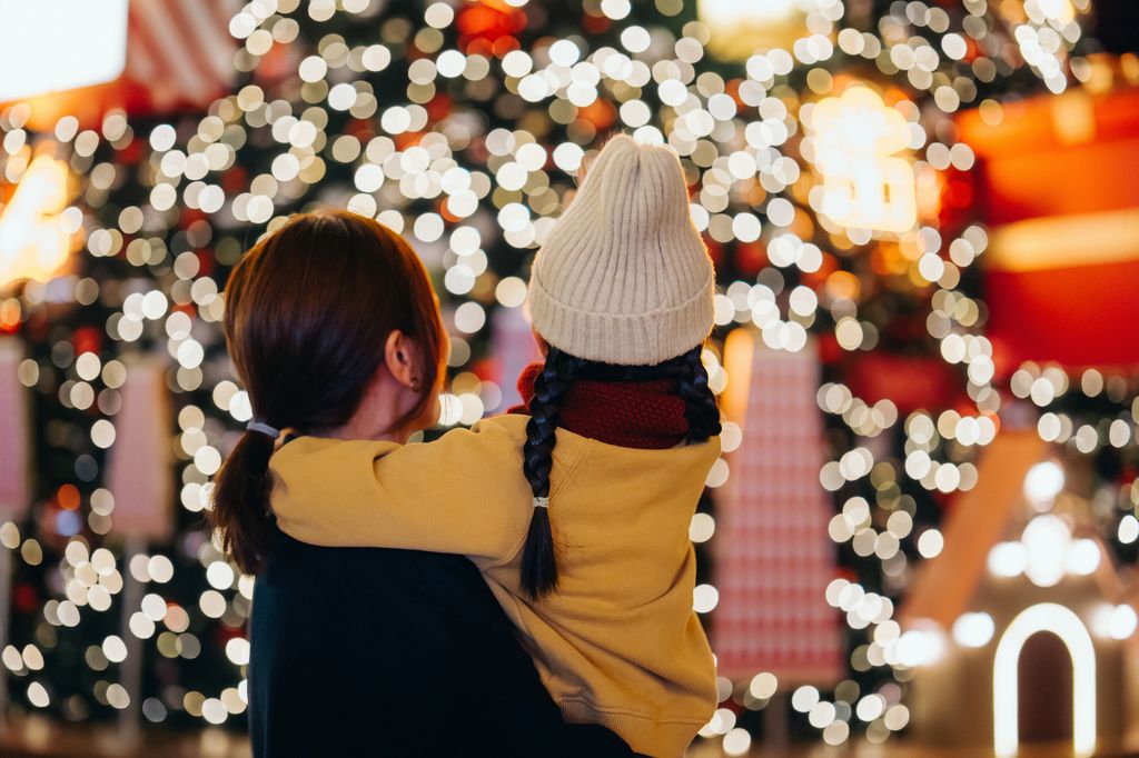 Rear view of young Asian mother carrying her lovely little daughter in arms, looking at illuminated Christmas tree in Christmas market. Family moment. Enjoying Christmas atmosphere. Holiday and festive vibes. Christmas is here
